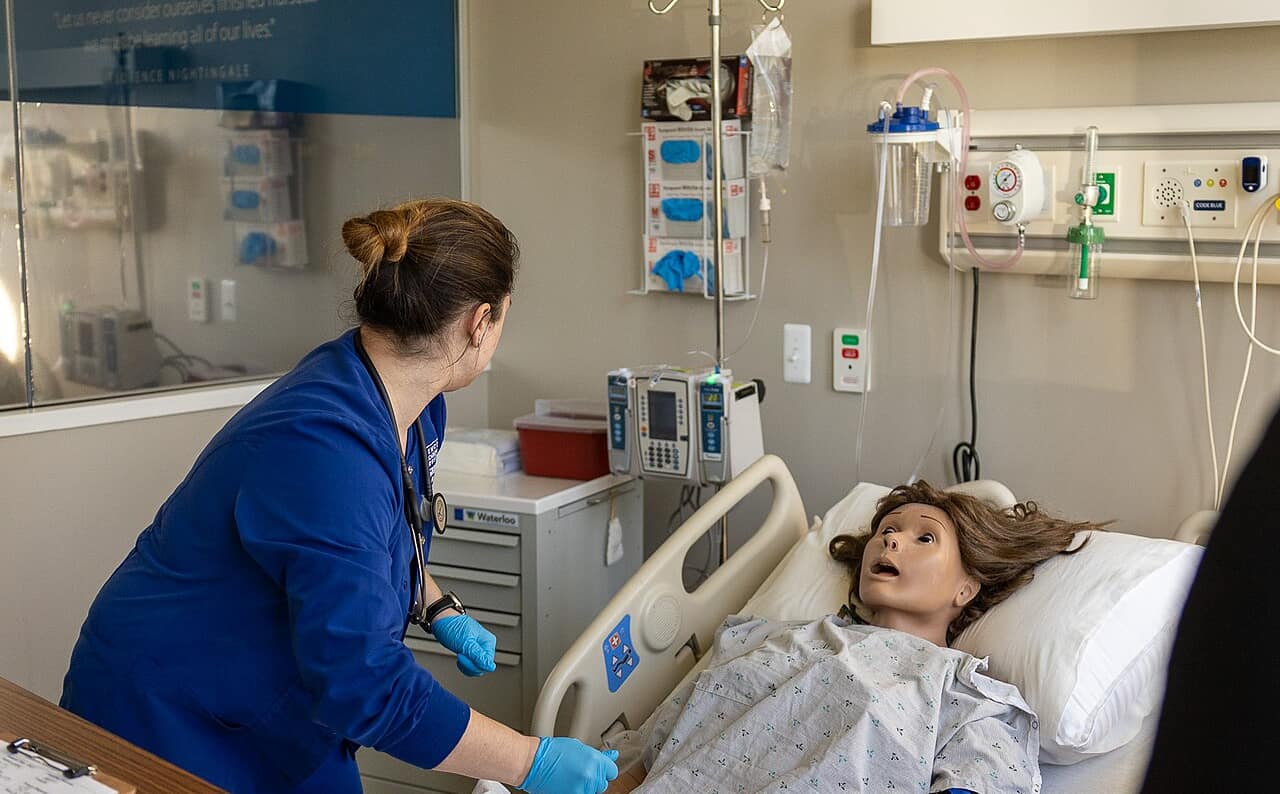 A nursing student in scrubs at a patient bedside in a simulation lab